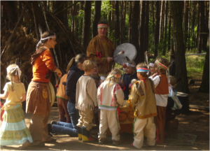 Kinder des Waldkindergarten Poppau stehen verkleidet in einem Kreis und halten sich an den Händen. Zwei Erzieher:innen stehen daneben und spielen Trommel.