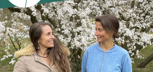 Simone und Alicia stehen vor einem blühenden Baum und lächseln sich an.